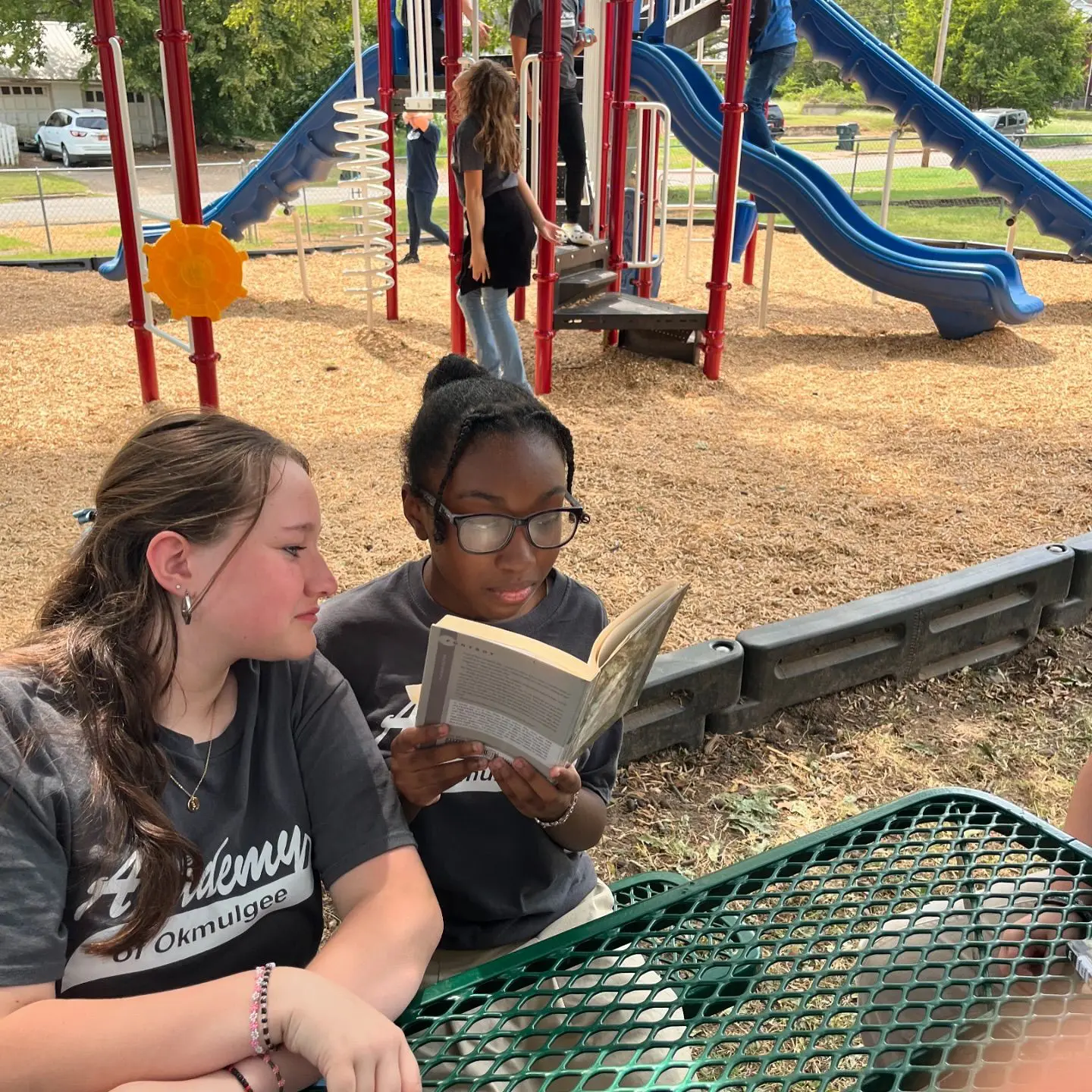 Two girls reading together at a playground picnic table.