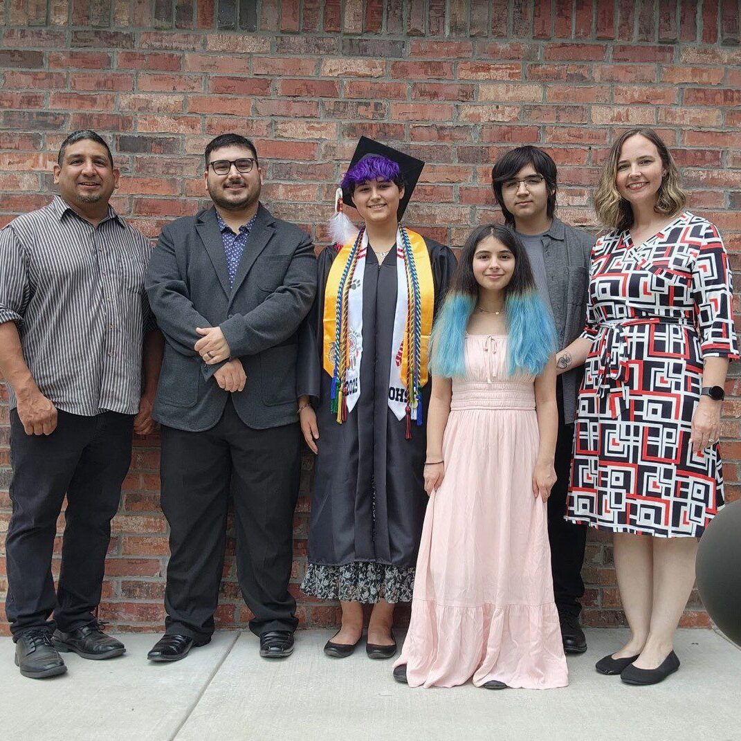 A graduate in cap and gown poses with family members outside a brick building.